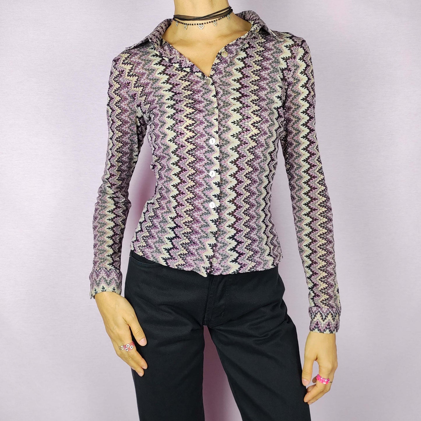 The Y2K Zigzag Mesh Top is a vintage 2000s shirt featuring a zigzag pattern in shades of black, pink, and beige. Made from a lightweight, stretchy mesh fabric, this button-up blouse offers a fitted silhouette with a pointed collar for a sleek look. Perfect for casual daytime wear.