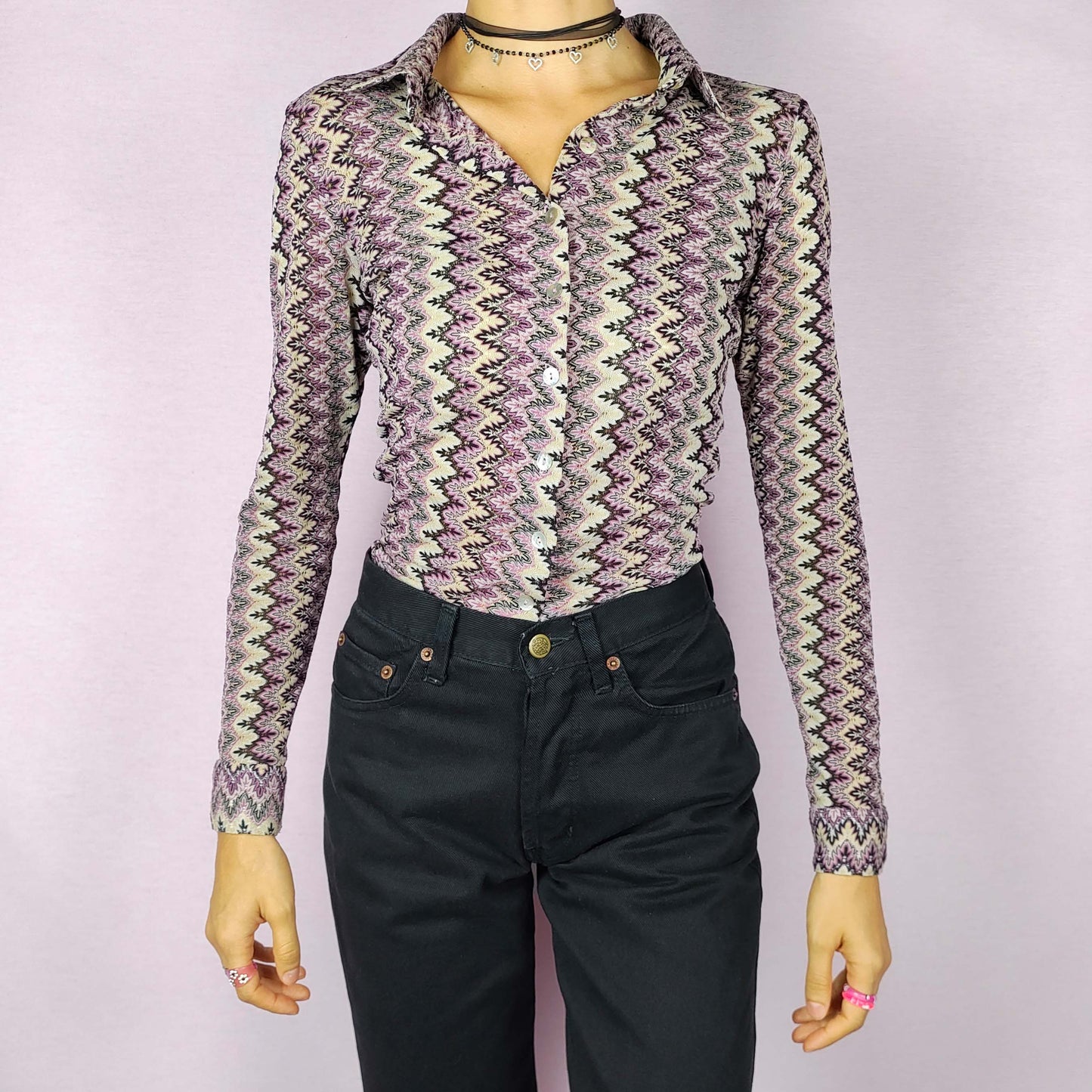 The Y2K Zigzag Mesh Top is a vintage 2000s shirt featuring a zigzag pattern in shades of black, pink, and beige. Made from a lightweight, stretchy mesh fabric, this button-up blouse offers a fitted silhouette with a pointed collar for a sleek look. Perfect for casual daytime wear.