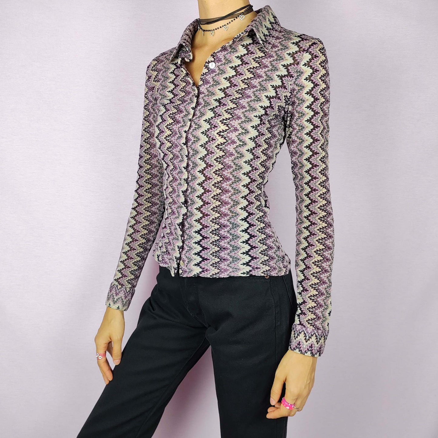 The Y2K Zigzag Mesh Top is a vintage 2000s shirt featuring a zigzag pattern in shades of black, pink, and beige. Made from a lightweight, stretchy mesh fabric, this button-up blouse offers a fitted silhouette with a pointed collar for a sleek look. Perfect for casual daytime wear.