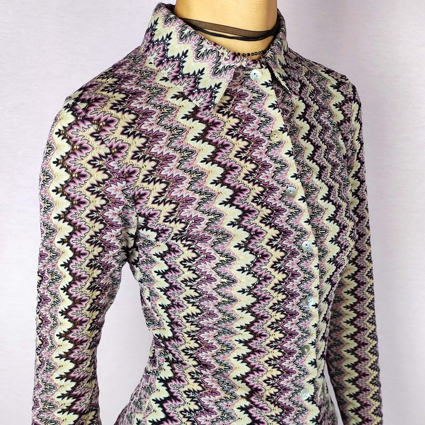 The Y2K Zigzag Mesh Top is a vintage 2000s shirt featuring a zigzag pattern in shades of black, pink, and beige. Made from a lightweight, stretchy mesh fabric, this button-up blouse offers a fitted silhouette with a pointed collar for a sleek look. Perfect for casual daytime wear.