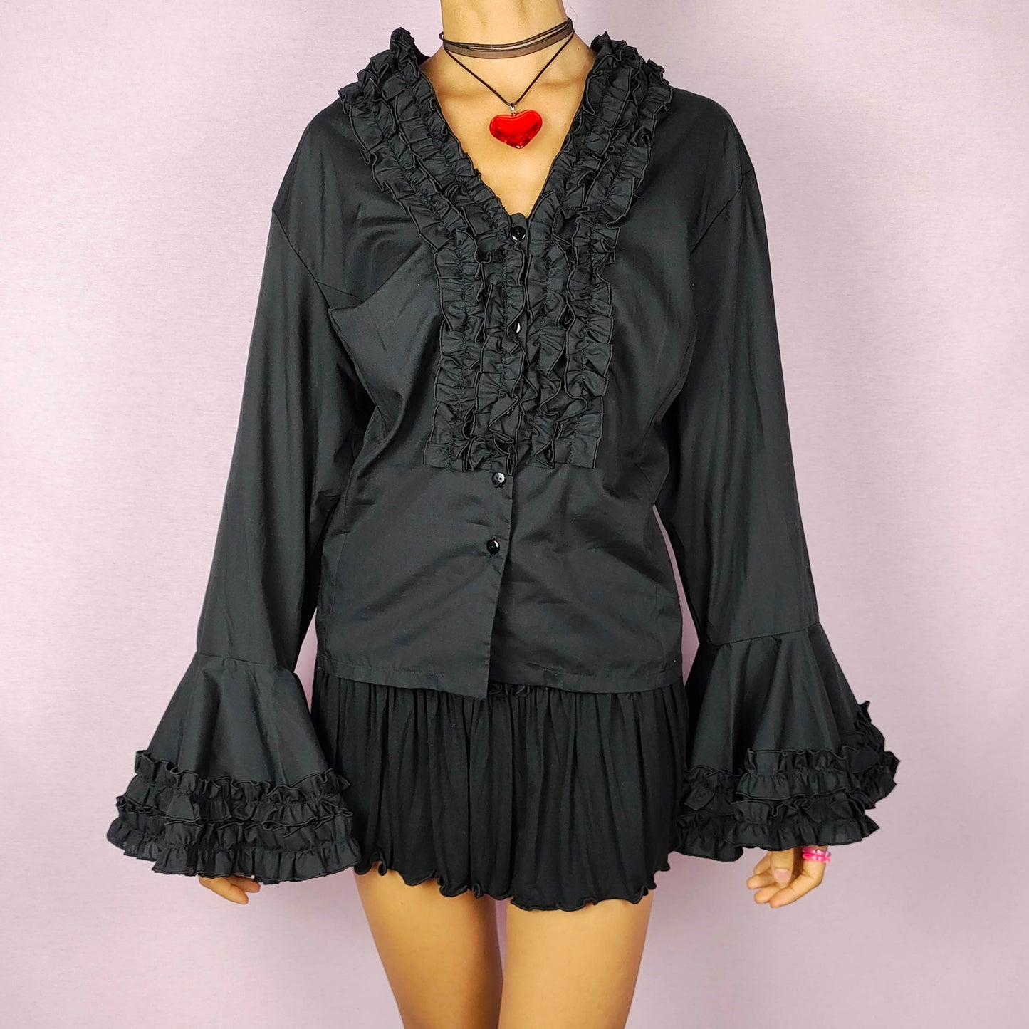 The Vintage 80s Black Ruffle Blouse is a statement shirt, featuring dramatic frill and ruffle detailing along the neckline and front button closure. This blouse includes voluminous bell sleeves with layered ruffles and frills. Perfect for a formal look or a night out.