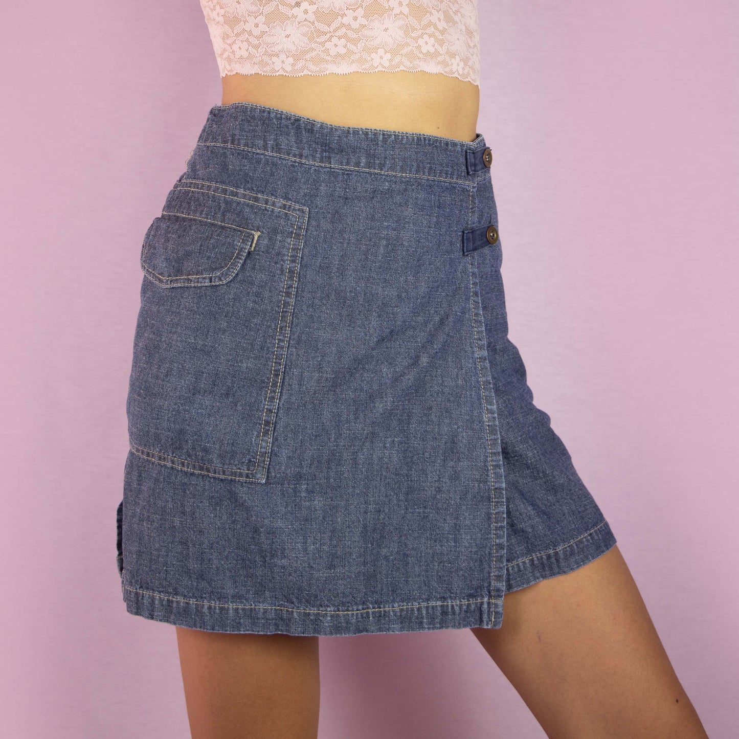 The Y2K Denim Wrap Mini Skirt is a vintage 2000s piece featuring a mid-rise fit and a unique wrap-style design with built-in shorts underneath. Made from medium-wash denim, the skirt has an asymmetrical front with button detailing and a side pocket. Ideal for casual wear and summer outings.