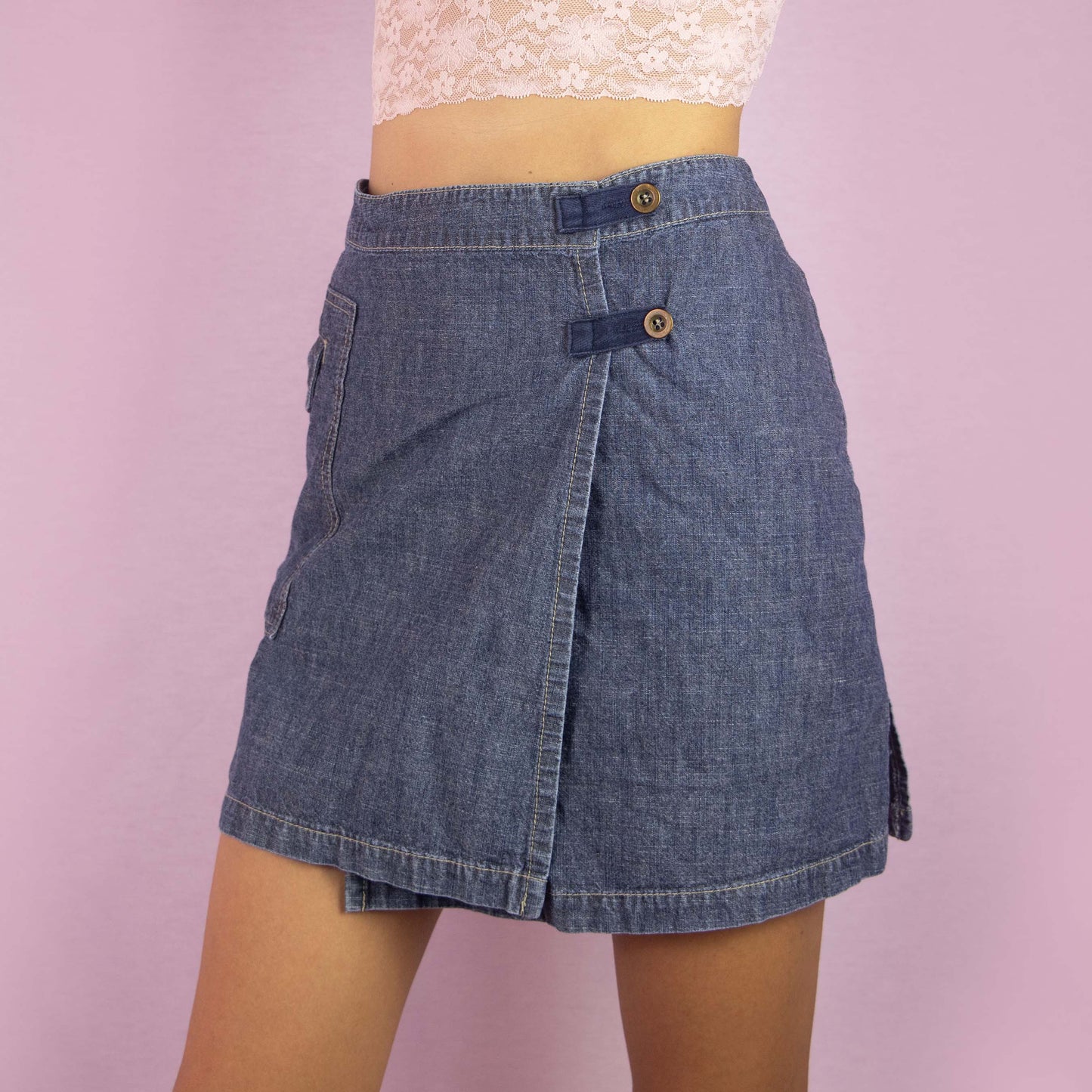 The Y2K Denim Wrap Mini Skirt is a vintage 2000s piece featuring a mid-rise fit and a unique wrap-style design with built-in shorts underneath. Made from medium-wash denim, the skirt has an asymmetrical front with button detailing and a side pocket. Ideal for casual wear and summer outings.