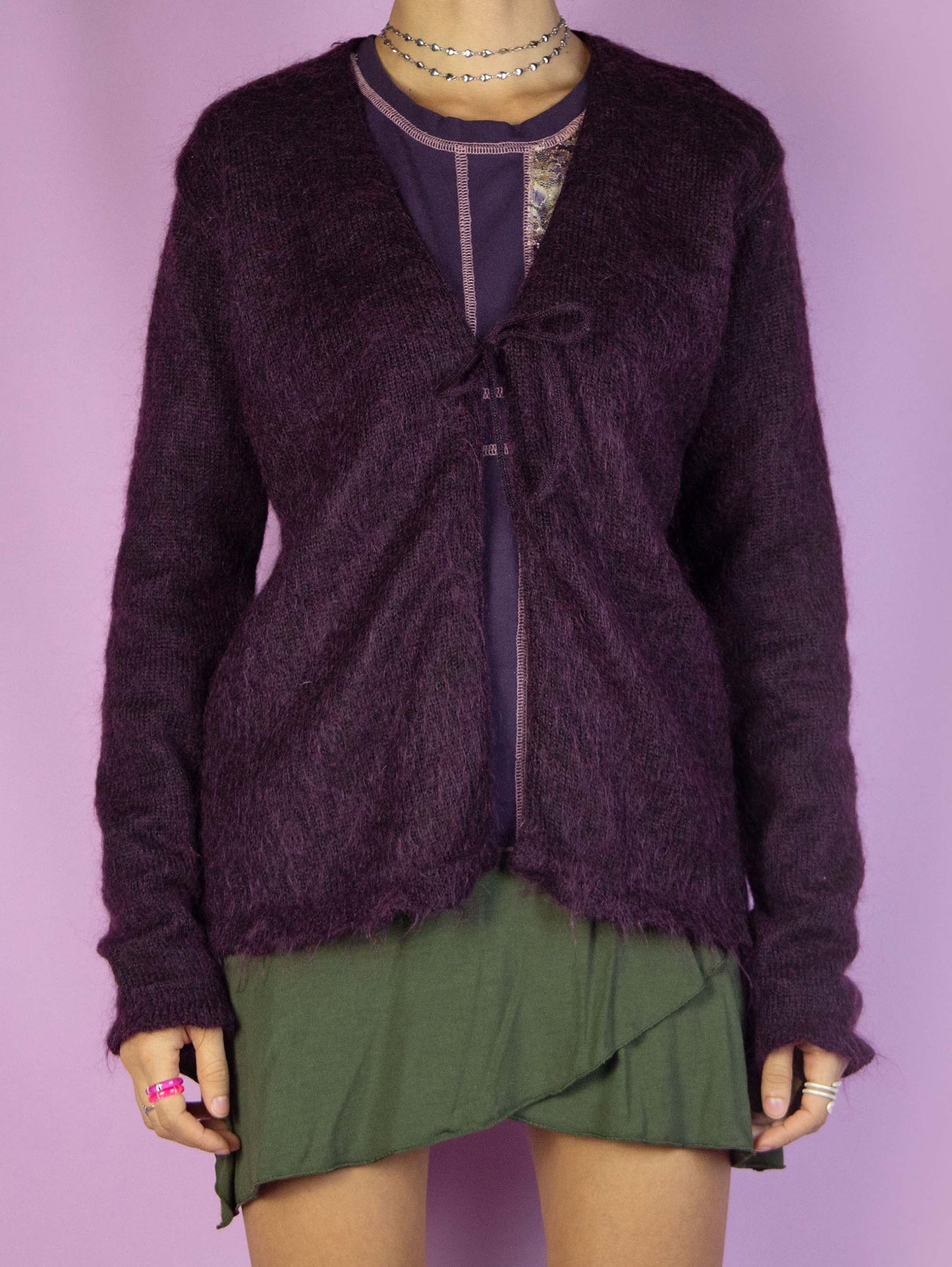 The Y2K Purple Tie Front Cardigan is a vintage 2000s sweater, crafted from a dark purple mohair blend. This cozy knit sweater features a tie front closure. Perfect for casual winter outfits.