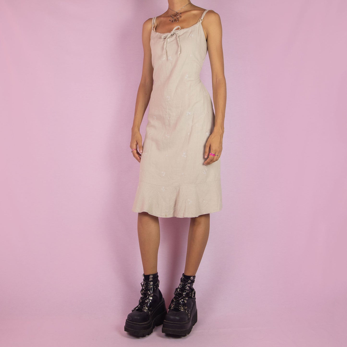 The Y2K Beige Linen Midi Dress is a vintage 2000s piece featuring a floral embroidery, tie-front detail, tie-back, and a back zipper closure. It also has adjustable spaghetti straps and a midi-length silhouette with a flared hem. Perfect for warm weather casual outings or a relaxed summer look.