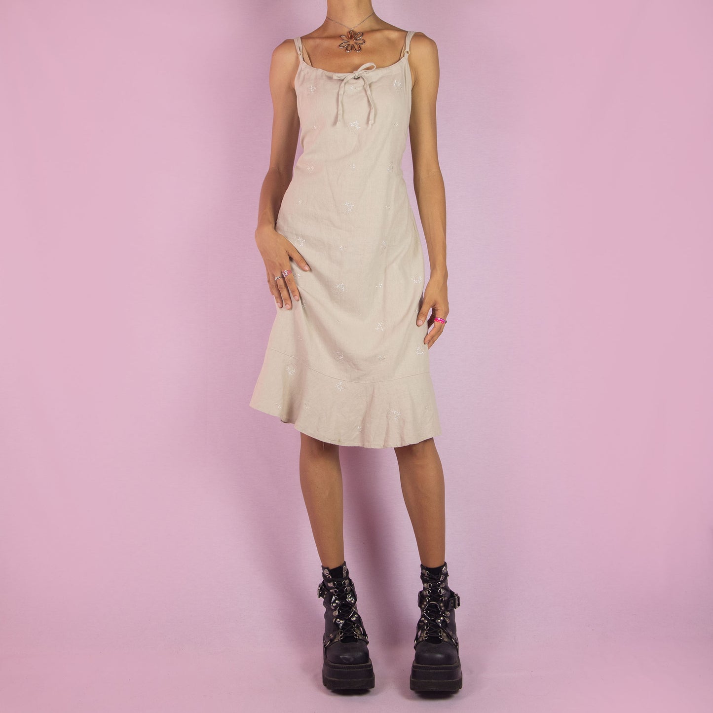 The Y2K Beige Linen Midi Dress is a vintage 2000s piece featuring a floral embroidery, tie-front detail, tie-back, and a back zipper closure. It also has adjustable spaghetti straps and a midi-length silhouette with a flared hem. Perfect for warm weather casual outings or a relaxed summer look.
