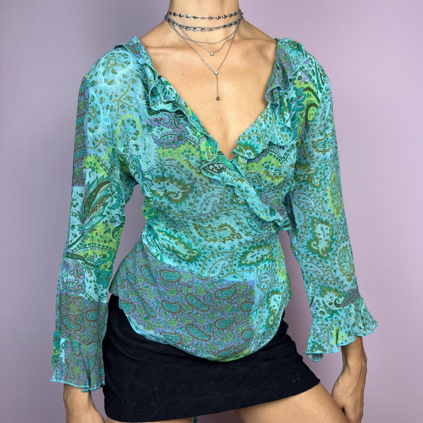 The Y2K Ruffle Wrap Tie Blouse is a vintage 2000s vibrant and playful summer statement piece, showcasing a lively green and blue paisley print with a semi-sheer finish. Featuring a flattering wrap-front design. adult world shop