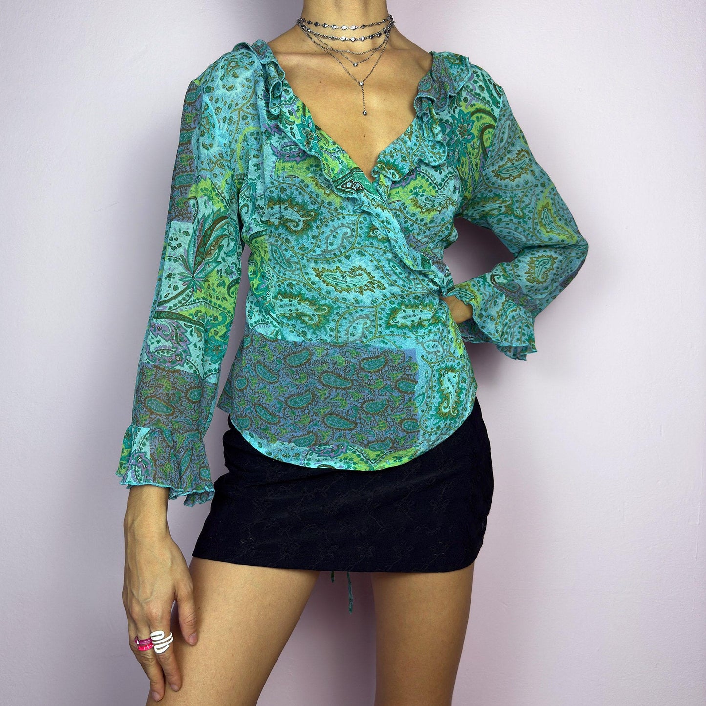 The Y2K Ruffle Wrap Tie Blouse is a vintage 2000s vibrant and playful summer statement piece, showcasing a lively green and blue paisley print with a semi-sheer finish. Featuring a flattering wrap-front design. adult world shop