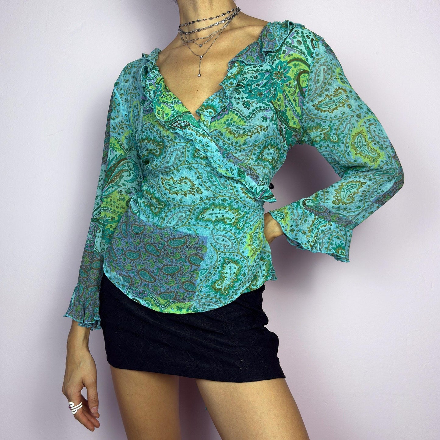 The Y2K Ruffle Wrap Tie Blouse is a vintage 2000s vibrant and playful summer statement piece, showcasing a lively green and blue paisley print with a semi-sheer finish. Featuring a flattering wrap-front design. adult world shop