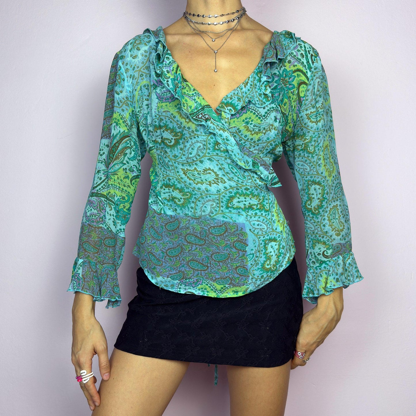 The Y2K Ruffle Wrap Tie Blouse is a vintage 2000s vibrant and playful summer statement piece, showcasing a lively green and blue paisley print with a semi-sheer finish. Featuring a flattering wrap-front design. adult world shop