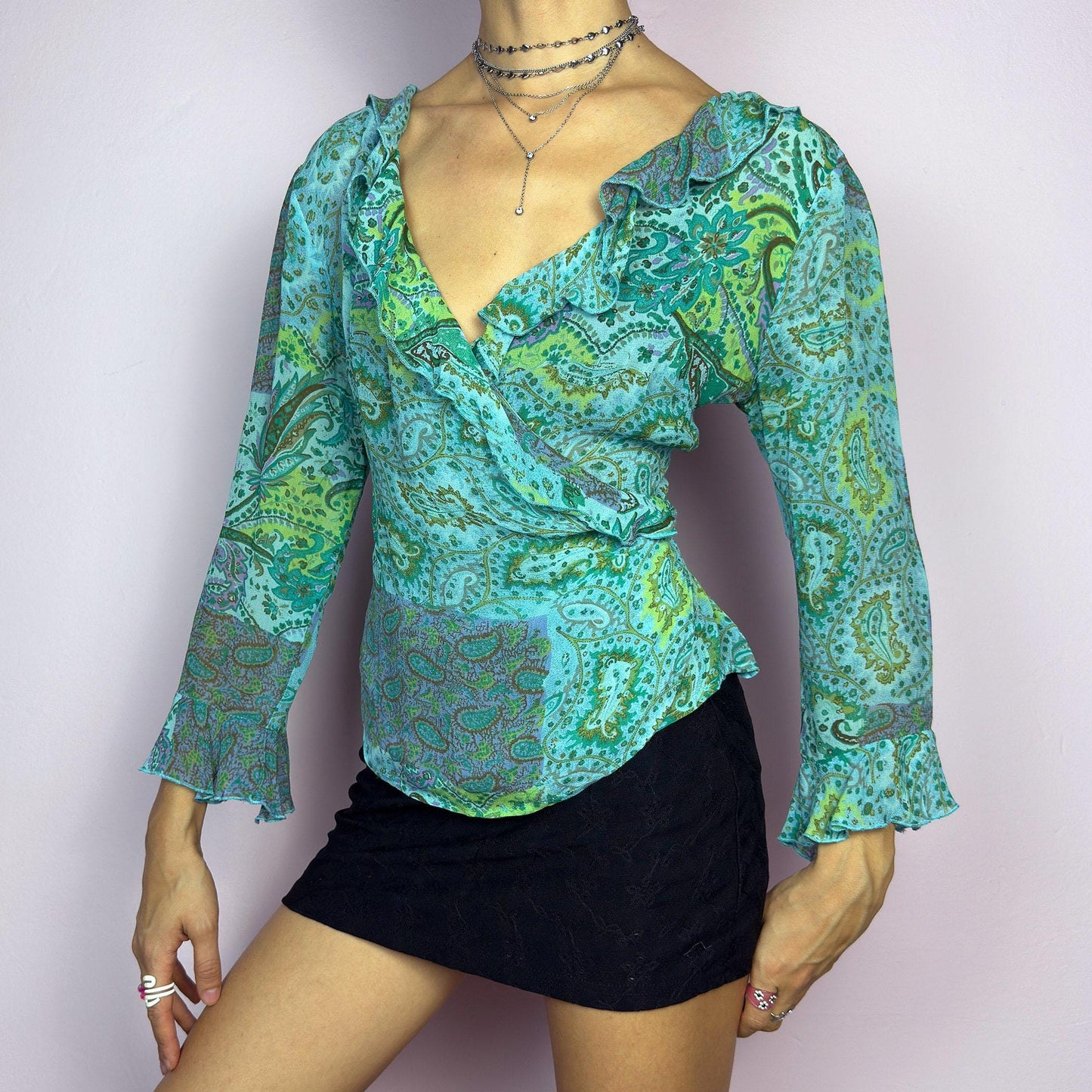 The Y2K Ruffle Wrap Tie Blouse is a vintage 2000s vibrant and playful summer statement piece, showcasing a lively green and blue paisley print with a semi-sheer finish. Featuring a flattering wrap-front design. adult world shop