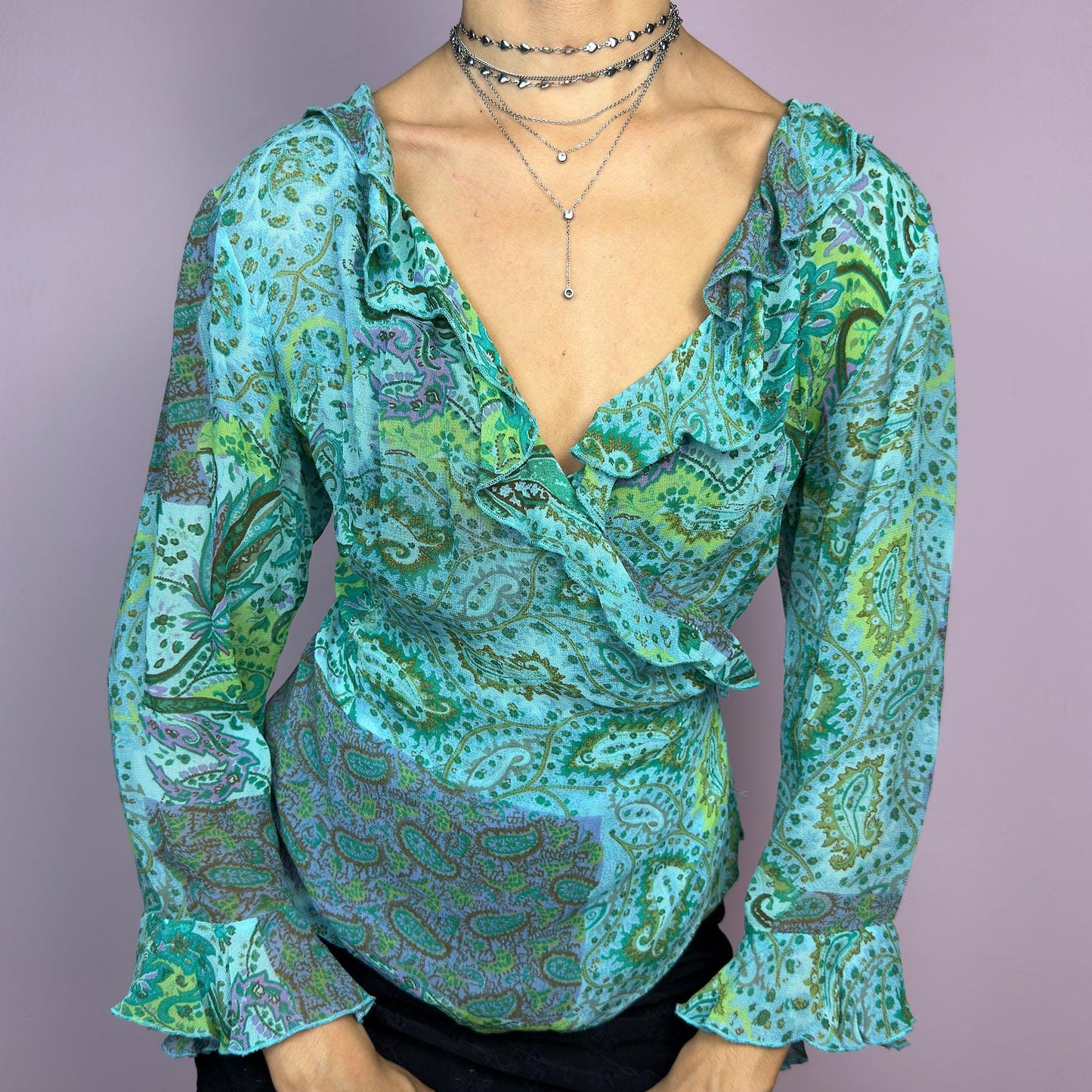The Y2K Ruffle Wrap Tie Blouse is a vintage 2000s vibrant and playful summer statement piece, showcasing a lively green and blue paisley print with a semi-sheer finish. Featuring a flattering wrap-front design. adult world shop