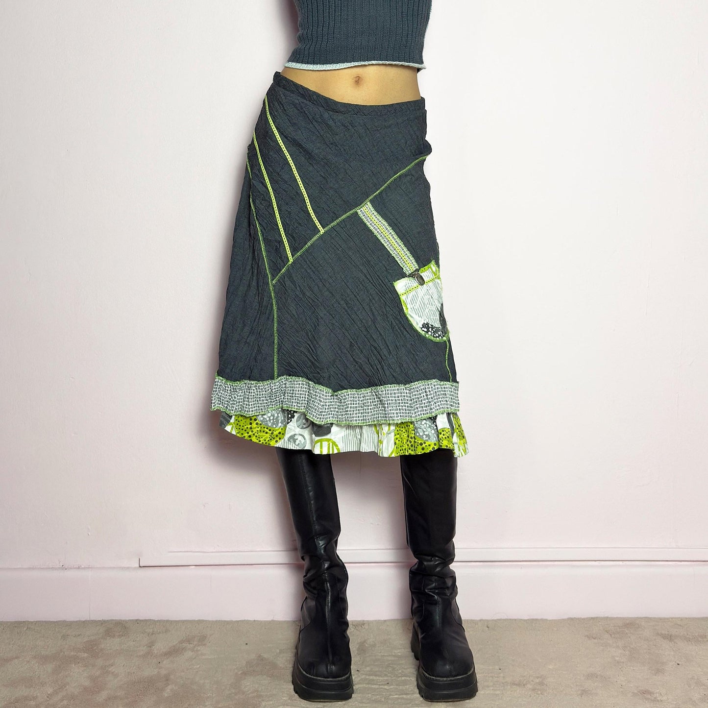 The Y2K Gray Patchwork Midi Skirt is a unique vintage 2000s piece featuring an eclectic mix of textures and contrasting neon green stitch details. Designed with a layered hem, ruffle trim, and a statement patch pocket with buckle detail, this skirt embodies a deconstructed boho aesthetic. The lightweight, crinkled fabric drapes effortlessly, while the elastic waistband ensures a comfortable fit, making it a perfect choice for bold, artsy outfits. Made in France. adult world shop