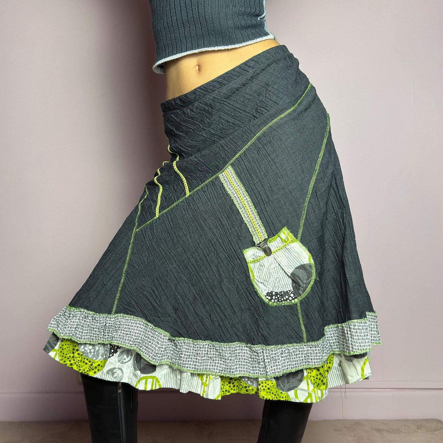 The Y2K Gray Patchwork Midi Skirt is a unique vintage 2000s piece featuring an eclectic mix of textures and contrasting neon green stitch details. Designed with a layered hem, ruffle trim, and a statement patch pocket with buckle detail, this skirt embodies a deconstructed boho aesthetic. The lightweight, crinkled fabric drapes effortlessly, while the elastic waistband ensures a comfortable fit, making it a perfect choice for bold, artsy outfits. Made in France. adult world shop