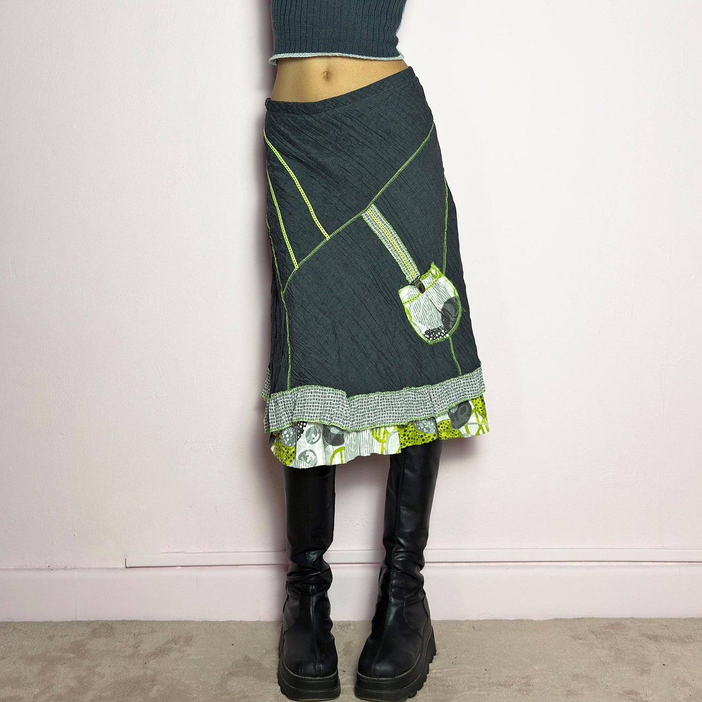 The Y2K Gray Patchwork Midi Skirt is a unique vintage 2000s piece featuring an eclectic mix of textures and contrasting neon green stitch details. Designed with a layered hem, ruffle trim, and a statement patch pocket with buckle detail, this skirt embodies a deconstructed boho aesthetic. The lightweight, crinkled fabric drapes effortlessly, while the elastic waistband ensures a comfortable fit, making it a perfect choice for bold, artsy outfits. Made in France. adult world shop
