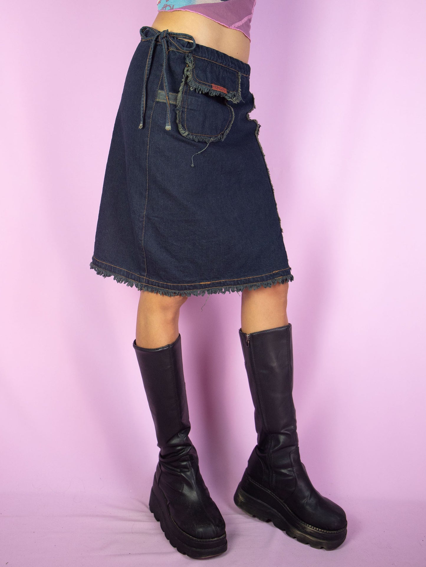 The Vintage Y2K Asymmetric Denim Mini Skirt is a dark denim navy blue skirt tied at the side with a pocket and frayed seam details. Cute cyber goth grunge gorpcore utility cargo deconstructed mini skirt circa 2000's.