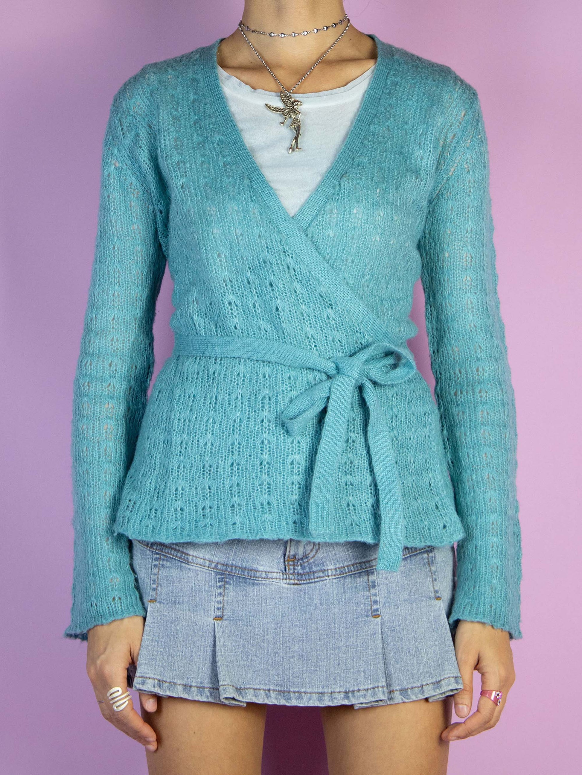 The Y2K Blue Wrap Cardigan is a vintage 2000s sweater that features a soft, lightweight knit in a turquoise shade. This wrap-style cardigan has a tie closure at the waist, creating a flattering silhouette, and long sleeves for added warmth.