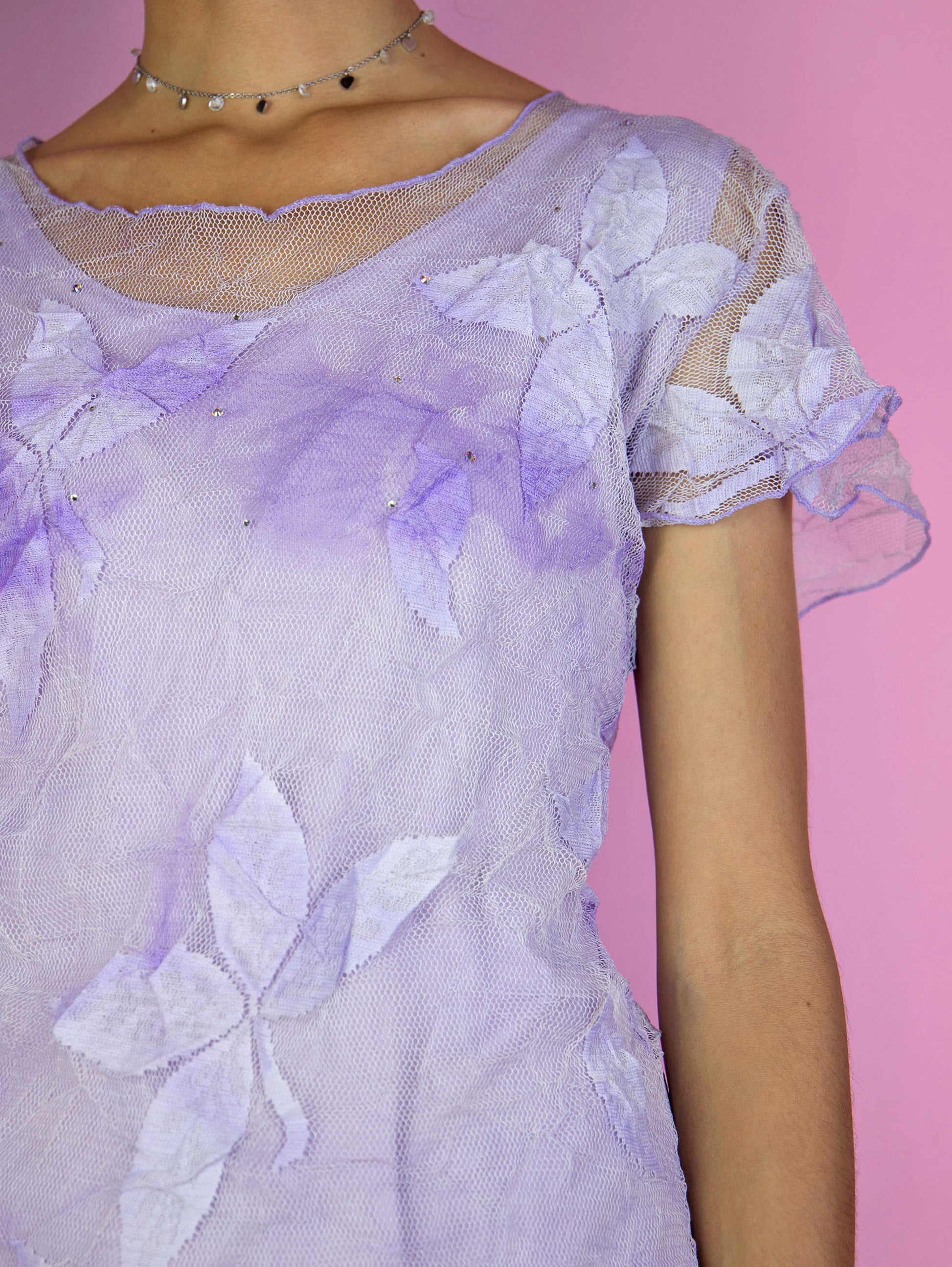The Y2K Lilac Mesh Sheer Top is a vintage short-sleeve, pastel purple semi-sheer mesh top adorned with delicate floral tulle and rhinestones. Romantic coquette 2000s fairy grunge shirt.