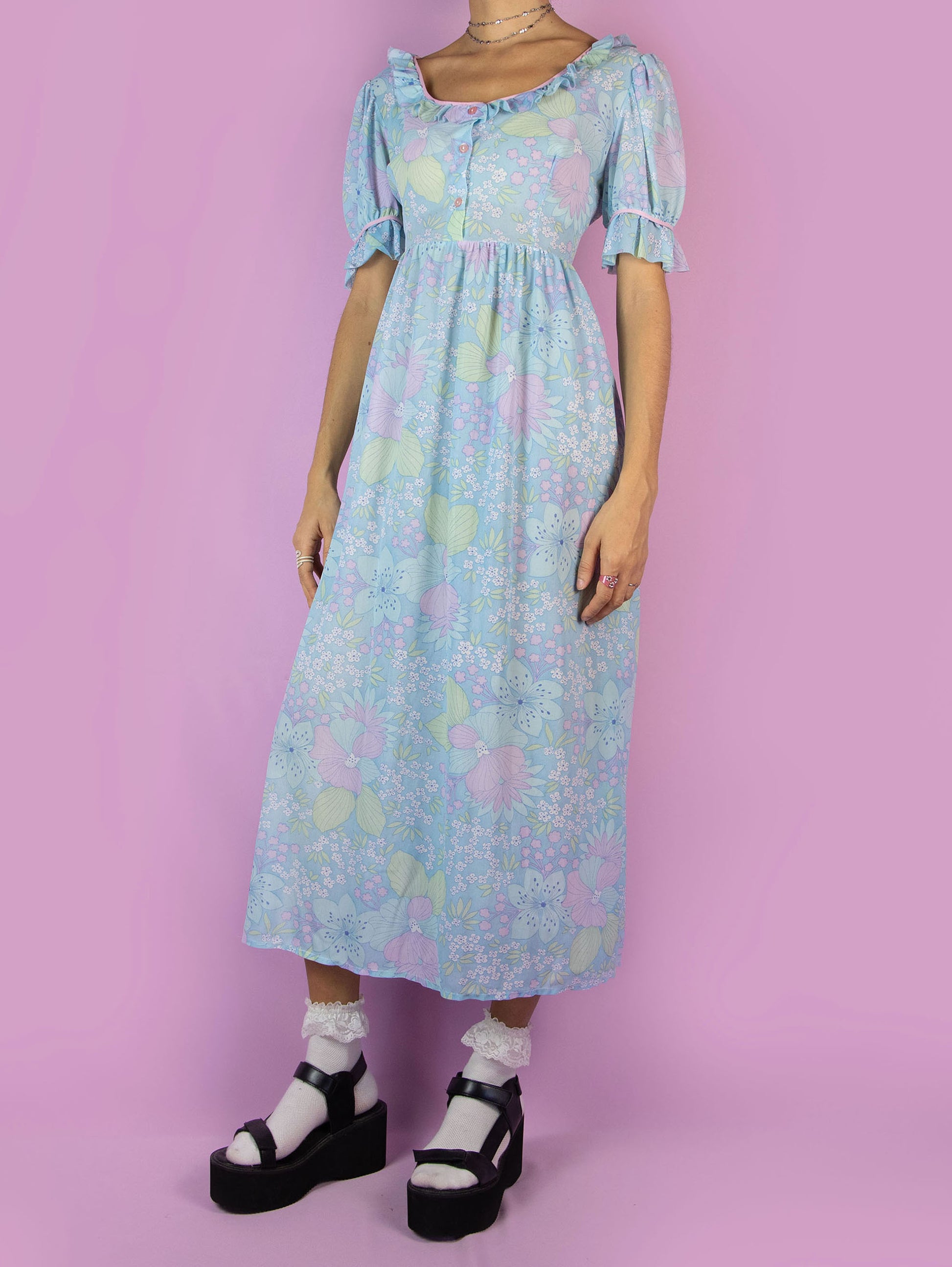 The Vintage 70s Floral Puff Sleeve Prairie Dress is a blue semi-sheer maxi dress in a multicolored floral pattern with short puff sleeves, ruffles, and buttons at the front. Country western inspired 1970s boho midi dress.