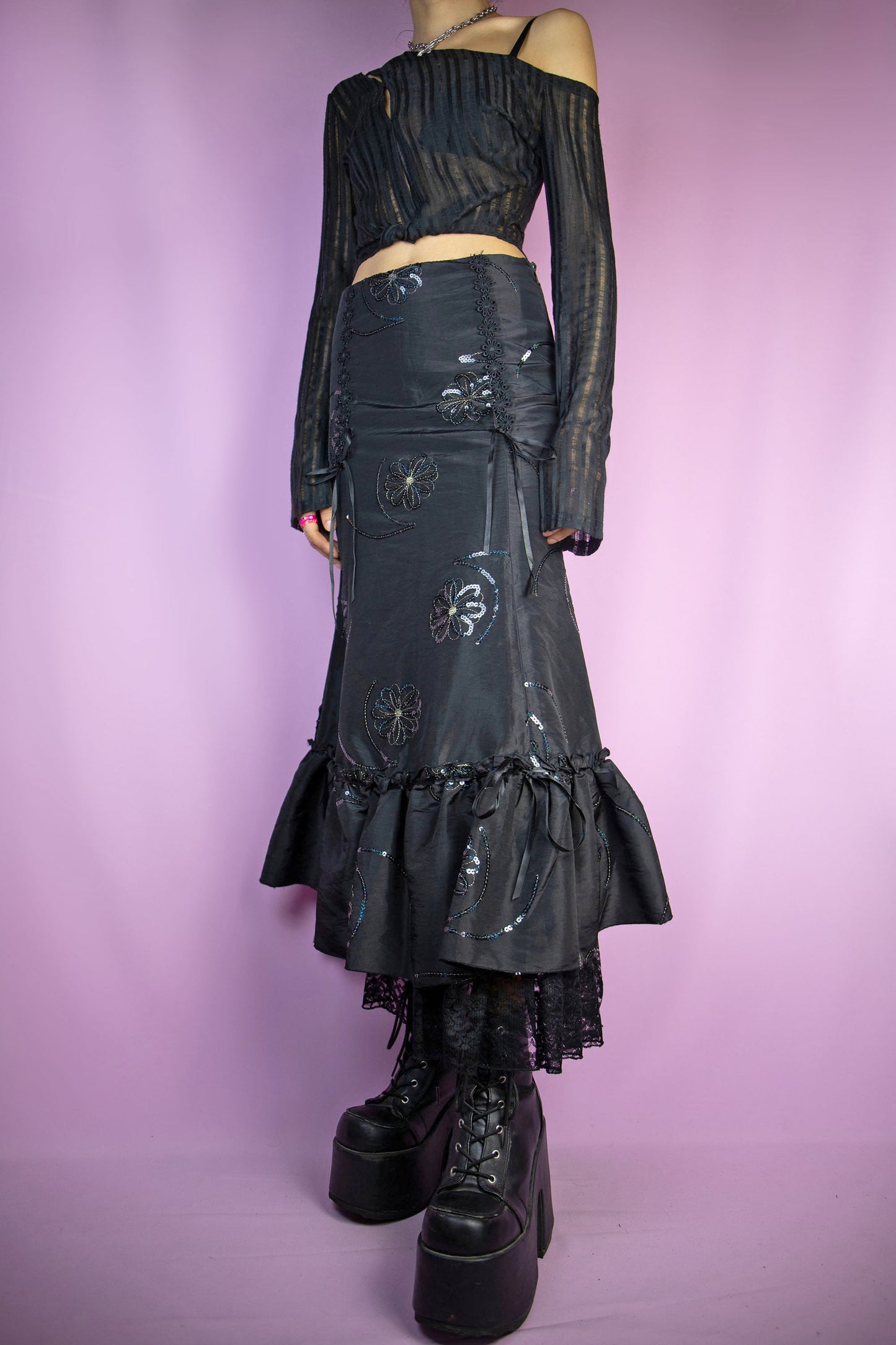The Vintage 90's Black Ruffle Maxi Skirt is an enchanting long black skirt adorned with sequin details, bows, a delightful ruffled hem, and a side zipper closure. This stunning fairy goth maxi skirt captures the essence of 1990s style.