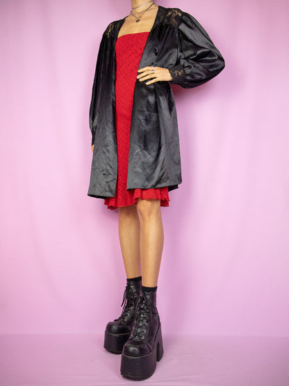 The Vintage 90's Black Satin Duster Jacket is a captivating romantic night goth peignoir from the 1990s. This shiny black balloon sleeve robe features lace details and a tie-up front, and it looks exceptionally cute when worn oversized.