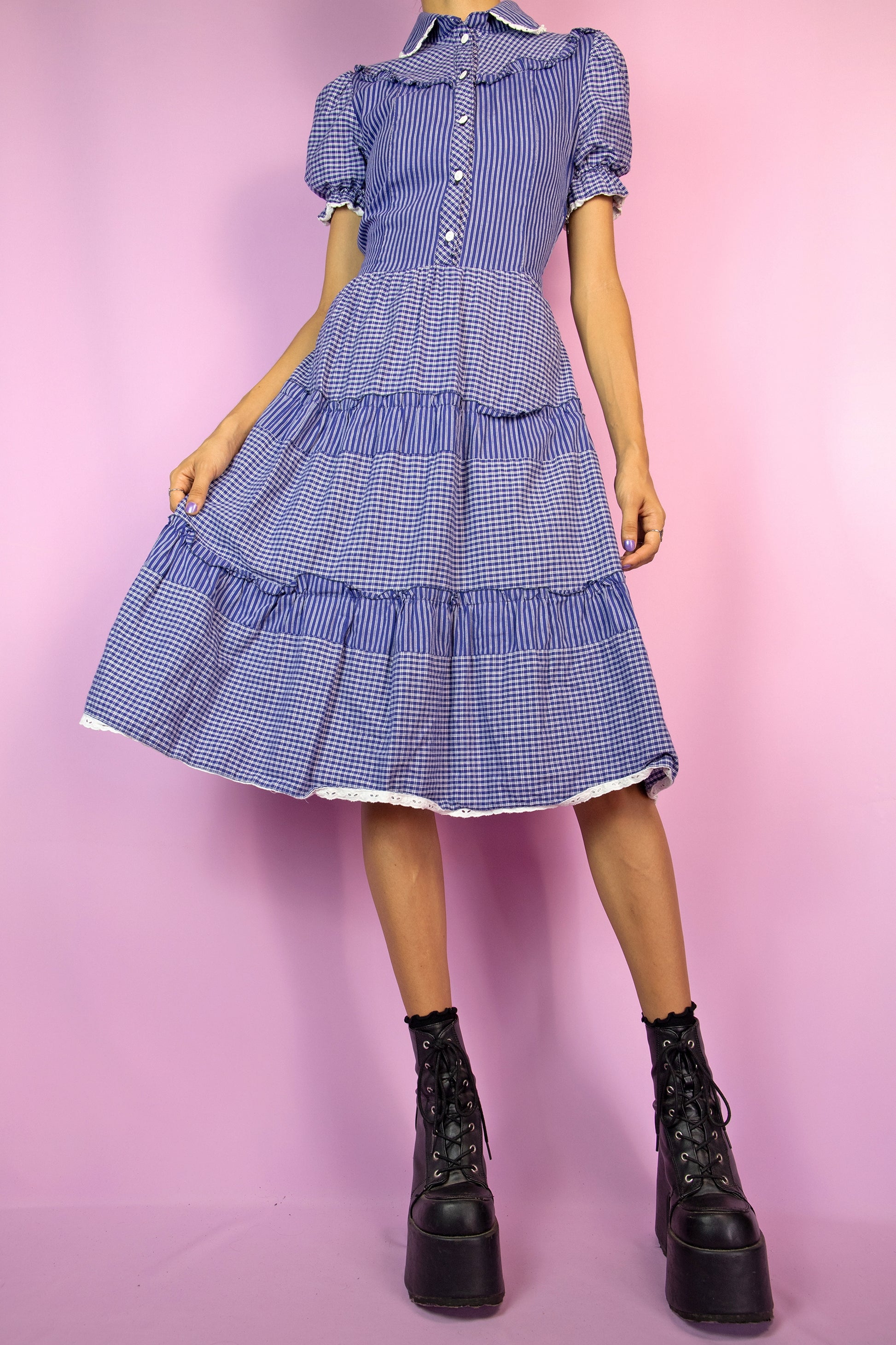The Vintage 80s Cottage Blue Check Dress is a blue and white striped and checkered tiered dress with puff sleeves, a collar, button front and ruffle details. It also has a side zipper closure and a matching fabric belt. Milkmaid inspired 1980s prairie plaid midi dress.