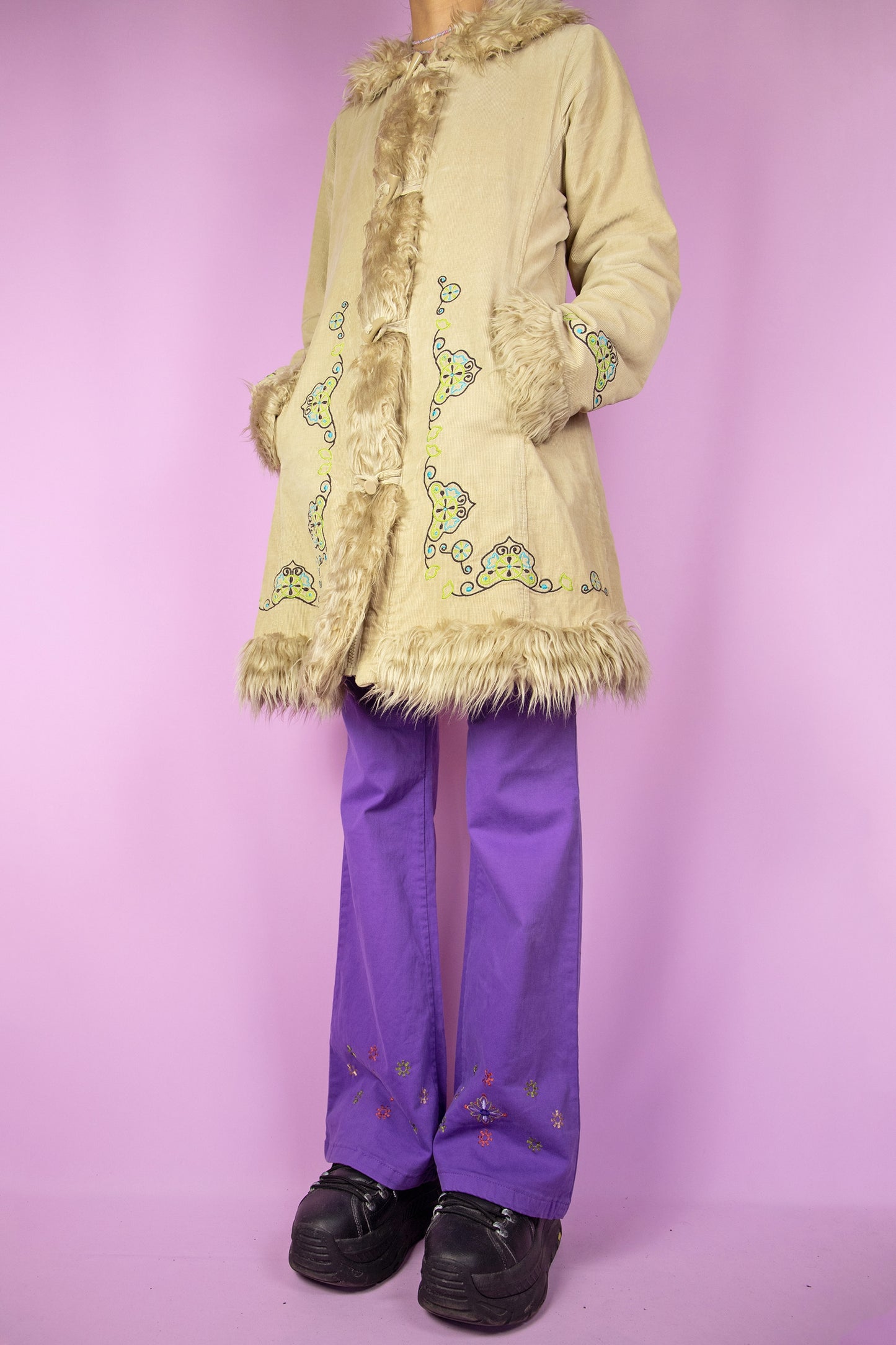 The Vintage Y2K Beige Afghan Inspired Coat is a light brown corduroy coat featuring a faux fur collar and cuffs, zip and button closure, pockets, and embroidered details. This iconic penny lane style winter coat from the 2000s serves as a boho fairy grunge statement jacket.