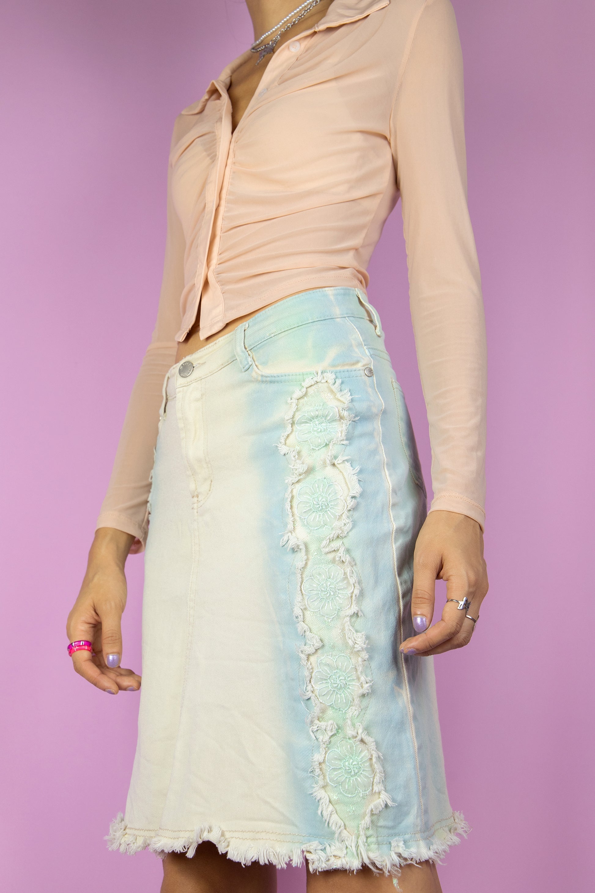 The Y2K Beige Denim Mini Skirt is a vintage beige and blue stretch skirt with frayed edges and floral appliqué details. Cyber 2000s jean mini skirt.