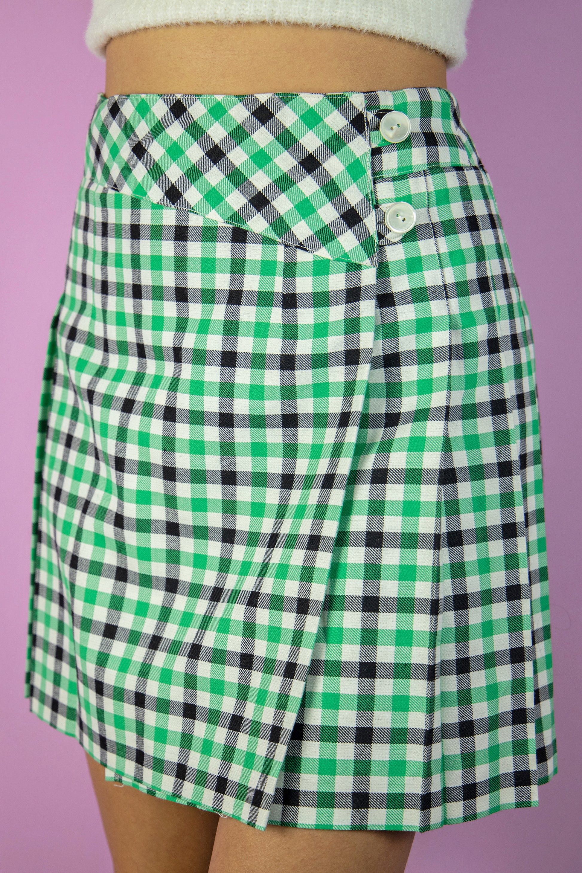 The Vintage 80s Check Pleated Mini Skirt is a classic preppy style green, black and white plaid pleated wrap mini skirt with side button closure.