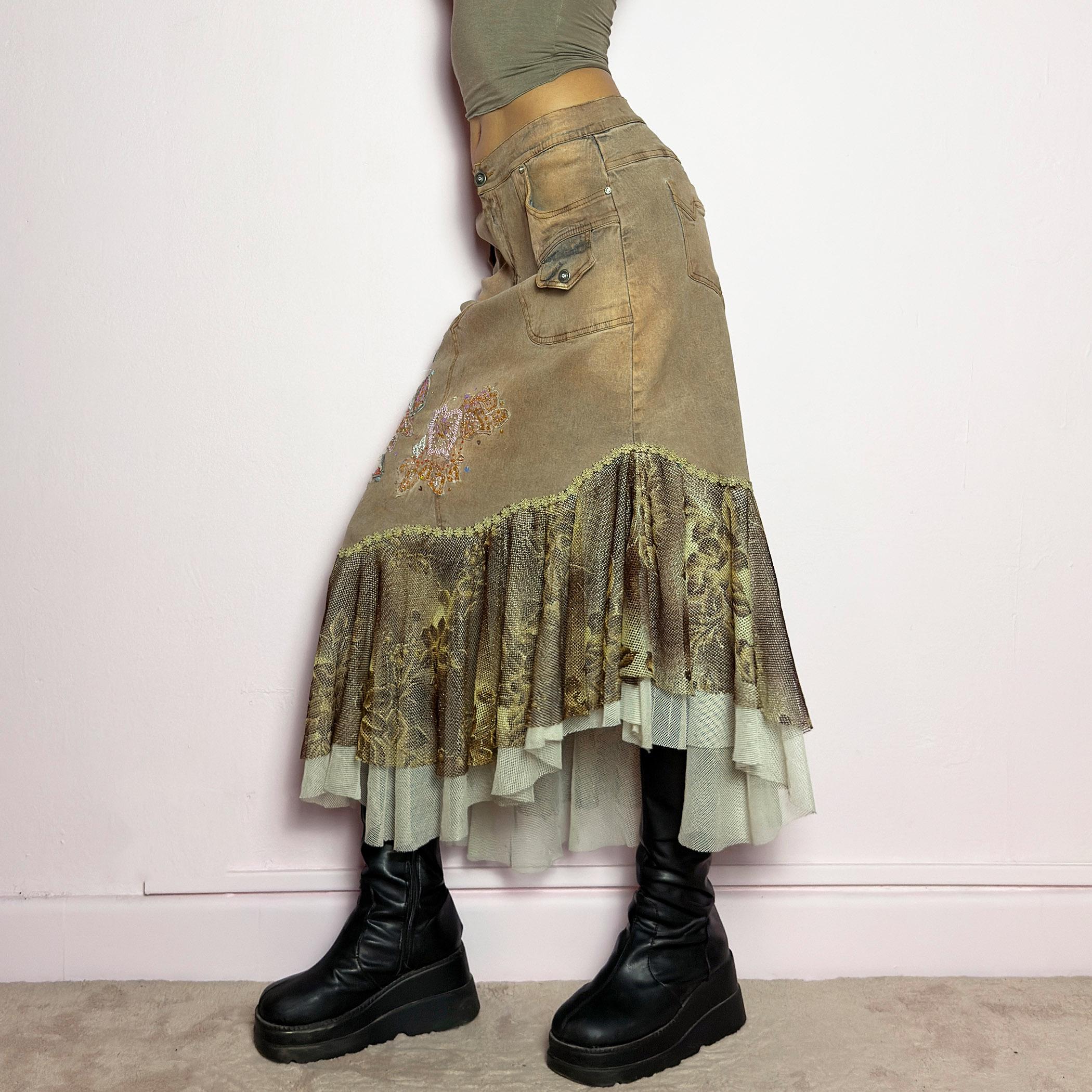 The Y2K Asymmetric Ruffle Midi Skirt is a vintage 2000s statement piece featuring a stretchy distressed tan denim base adorned with intricate beaded floral embroidery and sequin details. Designed with an asymmetric layered ruffle hem made of sheer lace and mesh with a metallic floral pattern, this skirt creates a dreamy, deconstructed aesthetic. It includes a button and zipper closure, belt loops, and pockets. adult world shop